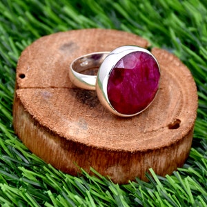 Può includere: Un anello d'argento con una grande pietra preziosa di rubino sfaccettato di forma ovale. L'anello è posto su una superficie di legno.