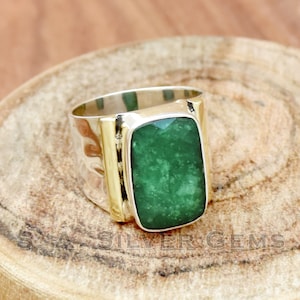 Può includere: Un anello in argento e oro con una grande pietra preziosa rettangolare verde. L'anello ha una fascia larga e una finitura testurizzata.