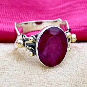 May include: A silver ring with a large, round, faceted ruby gemstone set in a silver bezel. The ring band is decorated with gold accents and small balls.