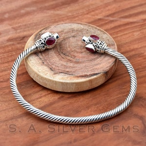 Könnte beinhalten: Ein silbernes Armreif-Armband mit einem gedrehten Design und zwei roten Edelstein-Akzenten. Das Armband ist offen und hat einen Scharnierverschluss.