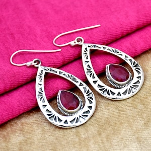 Peut inclure: Une paire de boucles d'oreilles pendantes en argent avec un design en forme de larme. Chaque boucle d'oreille présente une pierre précieuse rouge au centre.