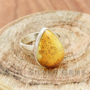 Pode incluir: Um anel em forma de lágrima com uma pedra de coral fossilizado amarelo engastada em prata de lei. A pedra tem um padrão detalhado de coral fossilizado.