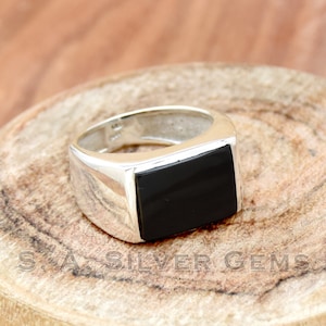 Puede incluir: Un anillo de plata con una piedra cuadrada negra. El anillo está sobre una superficie de madera.