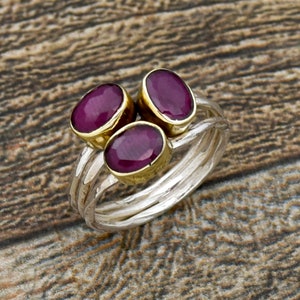 Könnte beinhalten: Ein silberner Ring mit drei ovalen Rubin-Edelsteinen, die in Gold gefasst sind. Der Ring hat eine strukturierte Band.