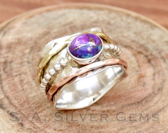 May include: A silver, gold, and copper spinning ring with a purple and blue gemstone. The ring is on a wooden surface.