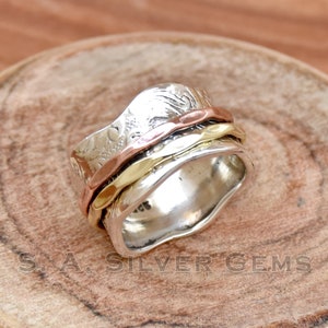 Può includere: Un anello girevole in argento, oro e rame con un designo testurizzato. L'anello è composto da tre bande che ruotano l'una intorno all'altra.