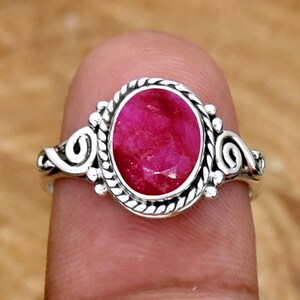 May include: A silver ring with a bright red ruby gemstone set in a decorative oval bezel. The ring has a twisted rope design on the bezel and ornate scrollwork on the band.