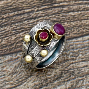 Puede incluir: Un anillo de plata y oro con tres bandas, que presenta un diseño floral con dos piedras preciosas rojas. El anillo tiene un acabado texturizado.