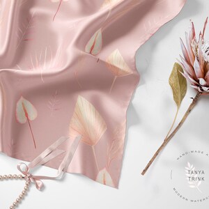 May include: A pink silk scarf with a delicate floral pattern featuring leaves and flowers. The scarf is draped over a white surface with a pearl necklace and a dried protea flower.
