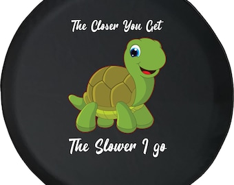 May include: Black spare wheel cover with a cartoon turtle in green and brown. The text "The Closer You Get The Slower I Go" is written in white above and below the turtle illustration.
