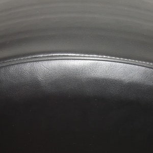 May include: Close-up of a black leather object with a curved shape. The leather has a textured surface and a stitched seam along the curved edge. The object appears to be a part of a larger item.