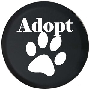 May include: A black circular cover featuring the word "Adopt" in white, above a white paw print. The cover is likely for a spare tire or similar item, promoting pet adoption.