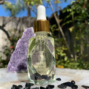Puede incluir: Una botella de vidrio transparente con una tapa dorada y un gotero llena de un líquido amarillo claro. La botella está etiquetada "Crystaline Corium Amethyst & Obsidian Dark Spot Elixir 10 ml/1 FL OZ". Piedras negras están esparcidas alrededor de la botella.