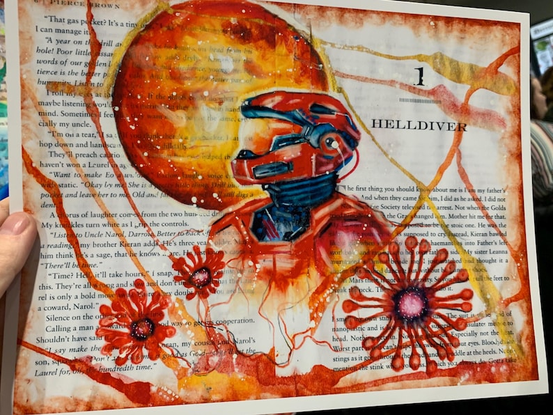May include: An artistic print on a book page featuring a red armored figure in a helmet, a large orange circular shape, and red floral-like patterns. The text "1 HELLDIVER" is printed in black ink above the figure, overlaid on printed text.