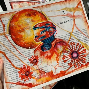 May include: An art print on a book page featuring a red armored figure in a helmet, a large orange planet, and stylized red floral shapes. The text '1 HELLDIVER' is visible at the top, with printed book text visible throughout the background.