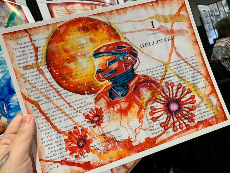 May include: An art print on a book page featuring a red armored figure in a helmet, a large orange planet, and stylized red floral shapes. The text '1 HELLDIVER' is visible at the top, with printed book text visible throughout the background.
