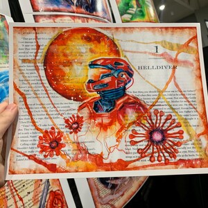 May include: A rectangular art print on a book page featuring a red armored figure in a helmet, a large orange circular shape, and red floral-like patterns. The text "1 HELLDIVER" is printed in black near the top right of the composition.
