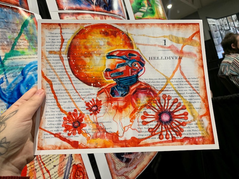 May include: A rectangular art print on a book page featuring a red armored figure in a helmet, a large orange circular shape, and red floral-like patterns. The text "1 HELLDIVER" is printed in black near the top right of the composition.