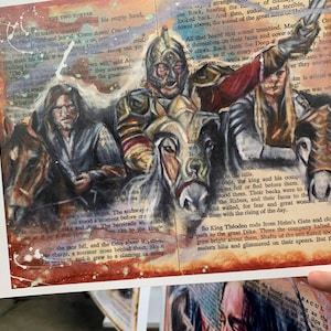 May include: Artwork featuring characters from a fantasy novel, painted on pages of a book. The illustration depicts several figures, including a warrior in armor, with horses and text from the book visible.