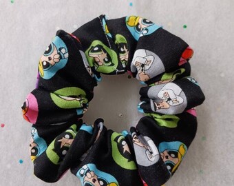 May include: Black fabric scrunchie with a repeating pattern of cartoon characters in green, blue, pink, and grey. The characters have large eyes and are set against a black background. The scrunchie is circular.