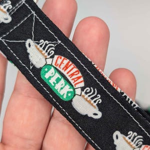 Może przedstawiać: Czarna smycz z tkaniny z białymi filiżankami do kawy i logo Central Perk w kolorze zielonym i czerwonym. Smycz ma srebrny metalowy zatrzask.