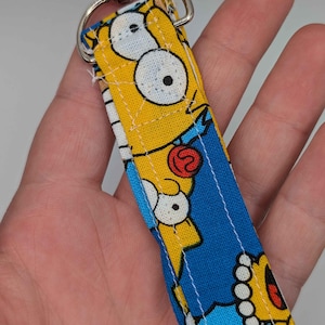 Może przedstawiać: Smycz z powtarzającym się wzorem twarzy postaci z kreskówki na żółtym tle z niebieskimi akcentami. Posiada srebrny metalowy zatrzask.