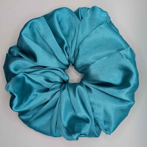 Peut inclure: Un grand chouchou en satin turquoise. L'accessoire pour cheveux est circulaire avec un design froncé, créant une apparence volumineuse. Le tissu a un léger éclat, reflétant la lumière. Le chouchou est de couleur unie et semble neuf.