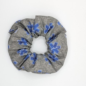 May include: A grey fabric hair scrunchie featuring a repeating pattern of blue six-pointed Star of Life medical symbols. The accessory has a gathered, circular design with visible fabric texture and consistent blue medical emblems throughout.
