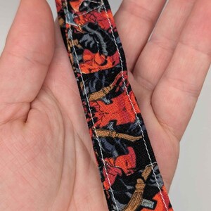 May include: A close-up of a patterned fabric strap with a silver metal loop. The fabric features a comic book-style design with red, black, and grey illustrations. White stitching outlines the edges of the strap.