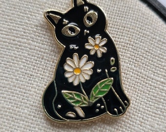 May include: A black cat enamel pin with gold-toned metal outlines. The cat features two white daisy flowers with yellow centers on its body, accompanied by green leaves. The cat has large, expressive eyes and pointed ears, rendered in a flat, graphic style.