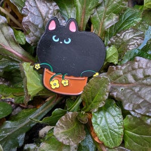 May include: A black cat enamel pin with pink ears and light blue eyes, sitting in a brown flower pot with yellow flowers. The pin is set against a backdrop of green and maroon leaves.