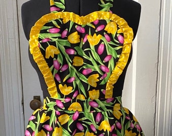 May include: A black apron with a heart-shaped top and a yellow ruffle trim. The apron is covered in a floral print of yellow and pink tulips on a black background.