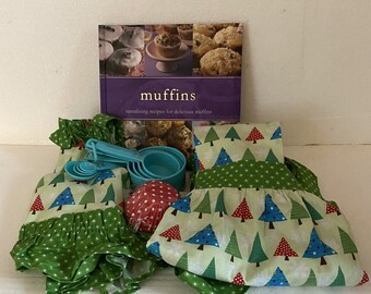 May include: A collection of baking supplies, including a cookbook titled "muffins," measuring cups and spoons, and a set of green and white patterned aprons and oven mitts. The aprons and oven mitts have a Christmas tree design. Red and white polka dot cupcake liners are also included.