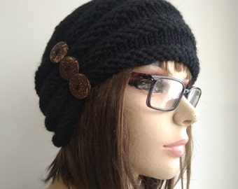 May include: A black knitted beanie with three brown coconut shell buttons. The beanie is worn by a person with brown hair and glasses.