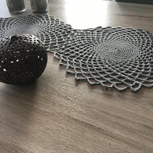 Könnte beinhalten: Eine graue gehäkelte Tischdecke mit einem runden Design liegt auf einem Holztisch. Im Vordergrund befindet sich eine braune Kokosnussschale mit Löchern.