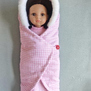 May include: A doll with dark brown hair and brown eyes wrapped in a pink and white gingham swaddle blanket. The swaddle features a white faux fur trim around the hood and is secured with a single small red button on the side of the fabric.