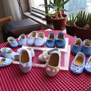 May include: A collection of baby shoes in various styles and patterns. The shoes include blue denim, striped, and floral designs. Some have bows or straps. The shoes are arranged on a red textured surface, with plants in the background.