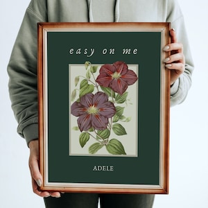 May include: Framed art print featuring a botanical illustration of two deep purple clematis flowers with green leaves. The print has the text "easy on me" at the top and "ADELE" at the bottom, set against a dark green background.