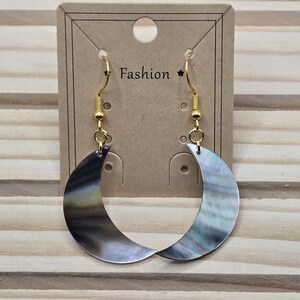 May include: A pair of crescent-shaped earrings with a mother-of-pearl finish. The earrings are suspended from gold-colored hooks and are displayed on a brown card with the word "Fashion". The earrings have a shimmering, iridescent quality.