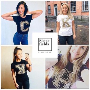 May include: Four women are wearing t-shirts with a large letter on the front. The letters are made of a leopard print fabric. The t-shirts are available in black and white. Sister fields clothing co.
