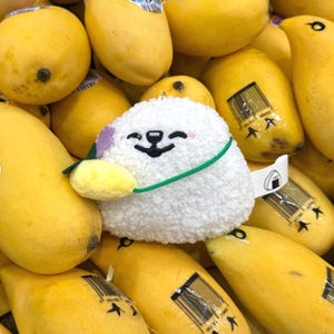 Puede incluir: Un personaje redondo y blanco de peluche con una cara sonriente, una correa verde y un pequeño accesorio de fruta amarilla, rodeado de mangos amarillos maduros. El personaje tiene una pequeña etiqueta con un gráfico.
