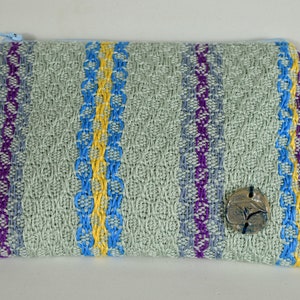 May include: A light green zippered pouch with a woven pattern in blue, purple, and yellow stripes. A small, round, ceramic button with a design is attached to the pouch.
