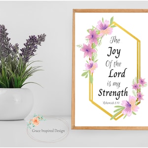 Puede incluir: Una impresión enmarcada con el texto "The Joy Of the Lord is my Strength" rodeada de flores moradas y rosas. La impresión está en un marco geométrico dorado. Un jarrón blanco con lavanda morada está a la izquierda.