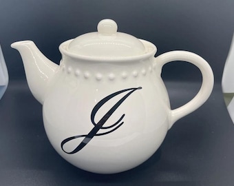 May include: A cream-coloured ceramic teapot featuring a black cursive letter "J". The teapot has a rounded body, a curved spout, and a loop handle. The lid is topped with a small knob. A row of small dots decorates the rim of the teapot.