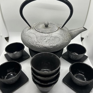 Puede incluir: Una tetera de hierro fundido plateada con asa y boquilla negras, rodeada de seis tazas y platillos de té negros. La tetera y las tazas tienen un diseño floral texturizado. El juego está sobre una superficie blanca.