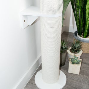 May include: White cat scratching post with a rope-covered pole and a white base. The post is mounted to a wall with a white bracket.