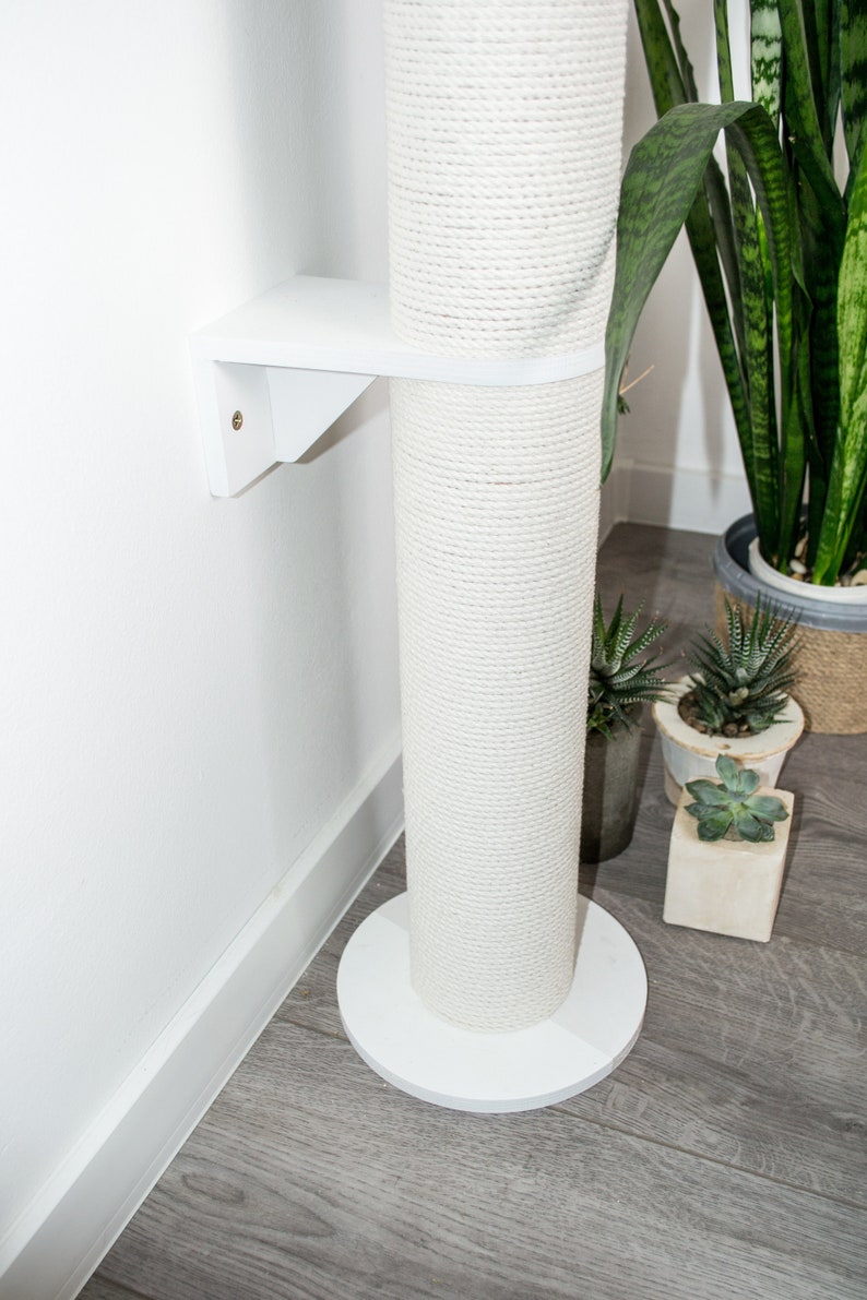 May include: White cat scratching post with a rope-covered pole and a white base. The post is mounted to a wall with a white bracket.