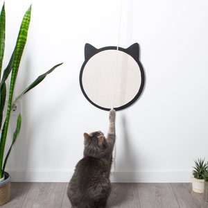 May include: A gray cat plays with a white rope hanging from a black and white cat-shaped scratching post mounted on a white wall. The scratching post is made of sisal rope and has a black outline of a cat's face. The cat is sitting on a gray wood floor.
