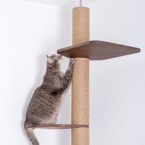 May include: A gray tabby cat climbing a wooden cat tree with two platforms and a sisal scratching post. The cat tree is attached to the ceiling. The cat is reaching for the top platform. The brand logo is visible.