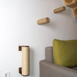 Pode incluir: Um poste de arranhar para gatos de madeira marrom com corda de sisal enrolada ao redor est&aacute; montado em uma parede branca. O poste est&aacute; ao lado de um sof&aacute; cinza com uma almofada verde.